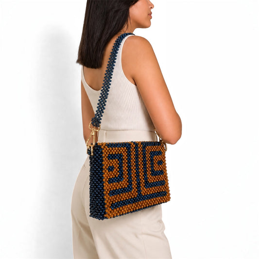 A woman wearing a white ribbed tank top and cream pants is shown from the side/back, wearing a beaded shoulder bag. The bag features a geometric pattern in dark navy blue and caramel-brown beads with gold-tone hardware.