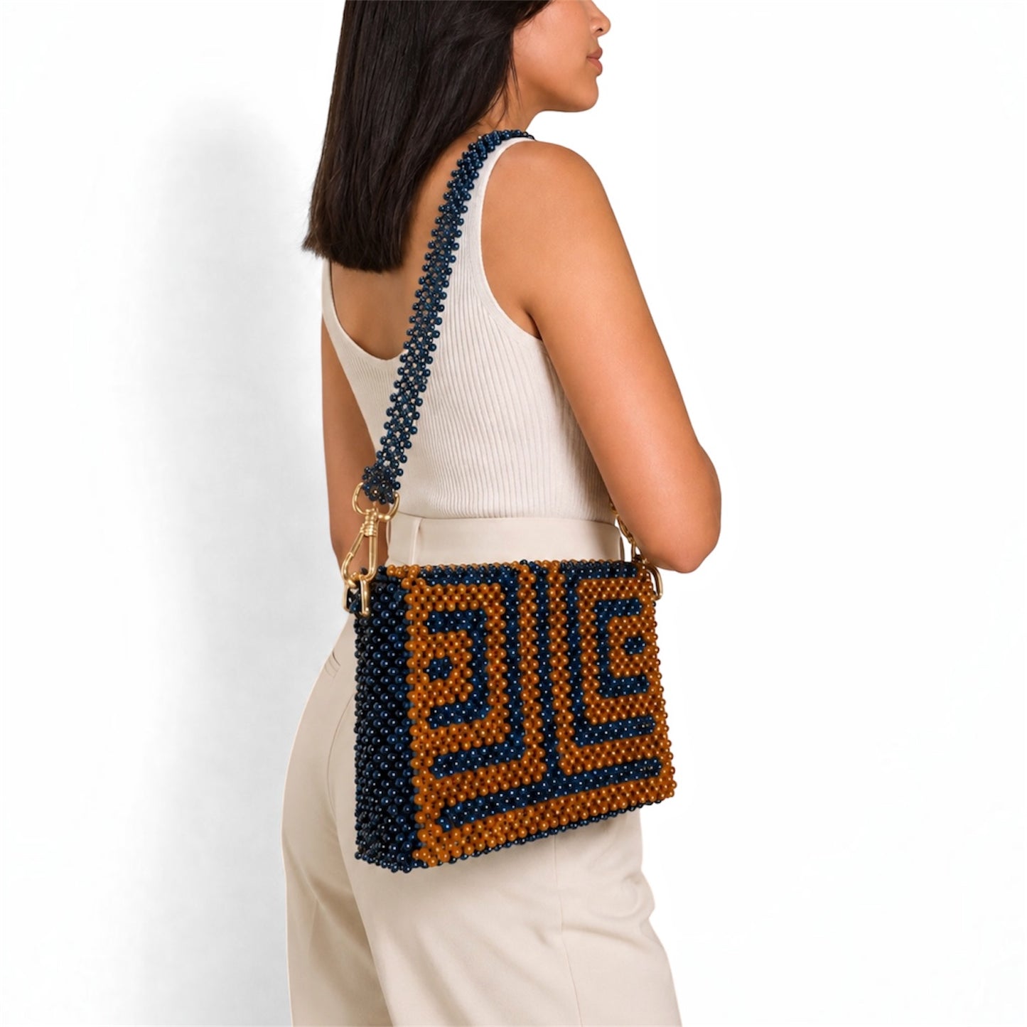 A woman wearing a white ribbed tank top and cream pants is shown from the side/back, wearing a beaded shoulder bag. The bag features a geometric pattern in dark navy blue and caramel-brown beads with gold-tone hardware.
